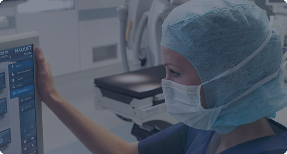 A healthcare worker in surgical scrubs and mask operating a touchscreen device in a medical facility