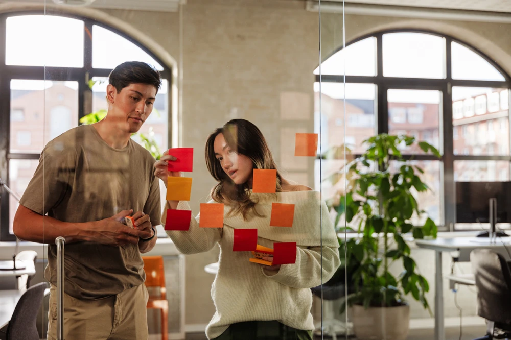 Man and woman placing a sticky note on a glass wall covered with other colorful sticky notes.